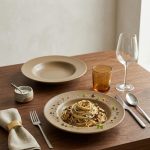 "Handmade ceramic dinner plates set on wooden dining table with pasta, wine glass and linen napkin — beige matte glaze artisan tableware India"